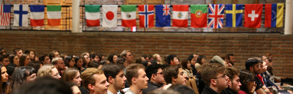 Grupo de estudiantes internacionales sentados en un salón. Al fondo se ven banderas de distintos países.
